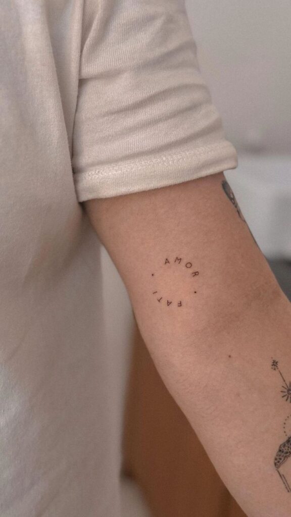 Minimalist arm tattoo reading amor fati in a circular pattern on a person's arm.