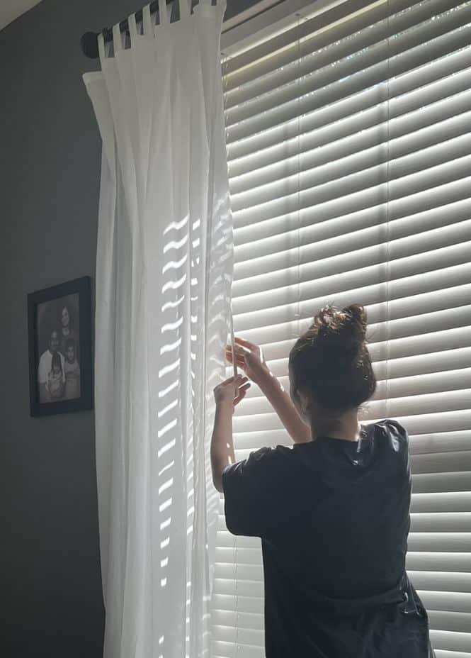 Person adjusting white curtains by a sunlit window with blinds in a cozy room.