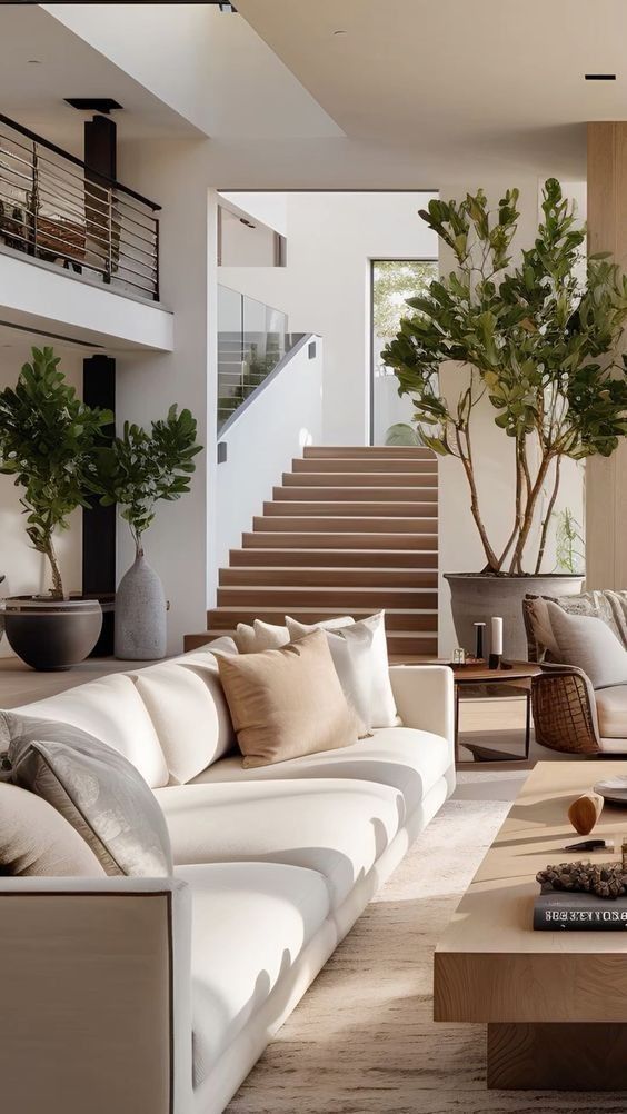 Modern living room with white sofa, neutral decor, potted plants, and staircase in a bright, open-concept space.