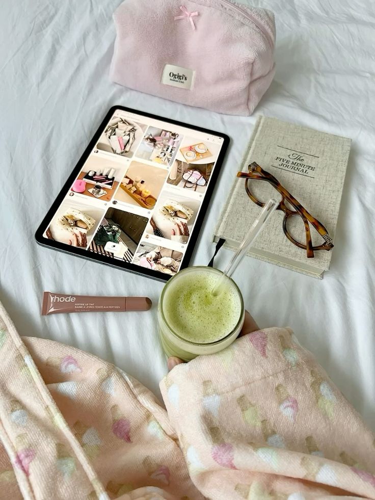 Cozy morning setup with tablet, journal, glasses, pink pouch, skincare, and green drink on a white bedspread.