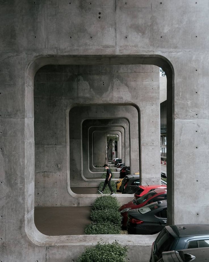 Concrete walkway with arches, cars parked beneath, and a person walking, creating a symmetrical urban scene.
