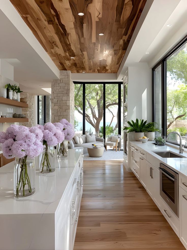 Modern kitchen with white cabinets, wood ceiling, large windows, and purple flowers on the counter, overlooking a garden.