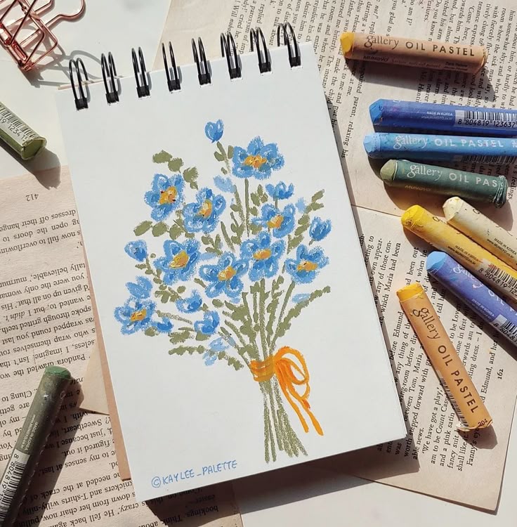 Sketchbook with blue floral drawing using oil pastels, surrounded by open books and colorful pastels.