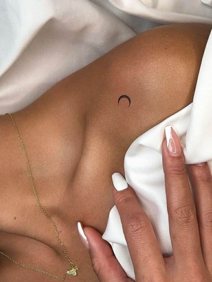 Minimalist crescent tattoo on skin, adorned with gold necklace and manicured nails, resting on white fabric.