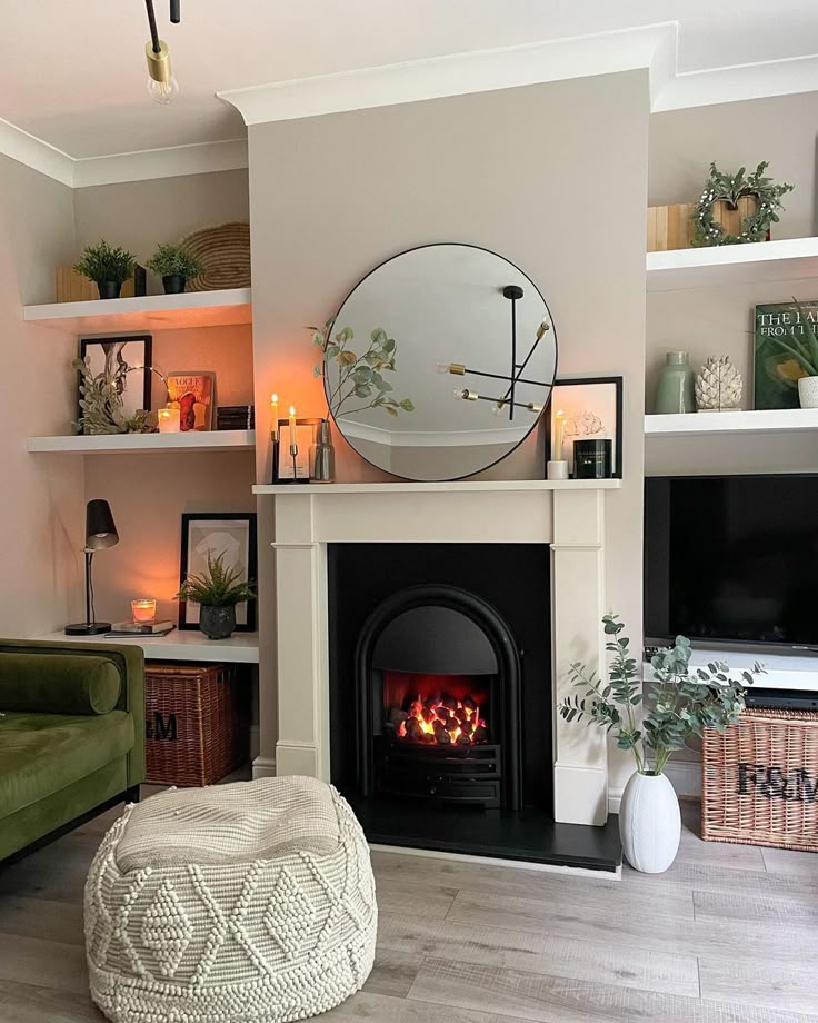 Cozy living room with a lit fireplace, round mirror, shelves, and green decor. Modern and inviting interior design.