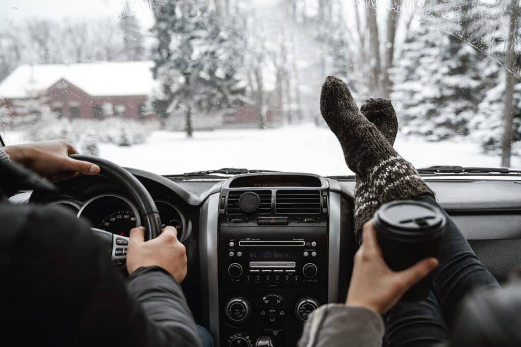 Winter road trip: relaxed passenger with cozy socks and coffee, snowy landscape ahead.