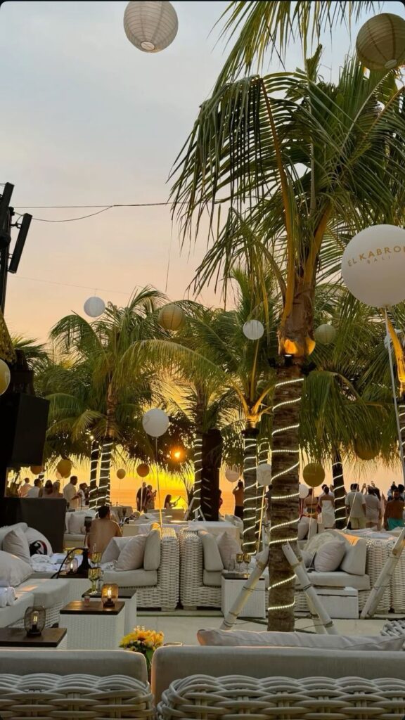 Luxurious beachside lounge with palm trees at sunset, white seating, and ambient lighting, perfect for a tropical getaway.
