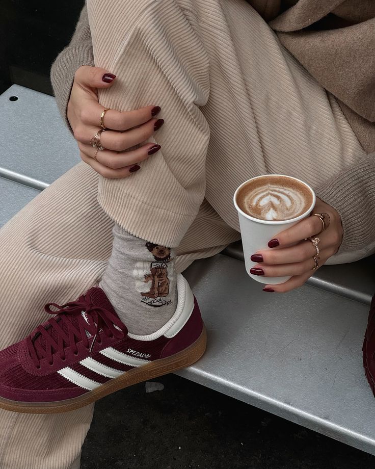 Woman with maroon sneakers and coffee, wearing cozy socks and beige pants, holding cup with latte art in outdoor setting.