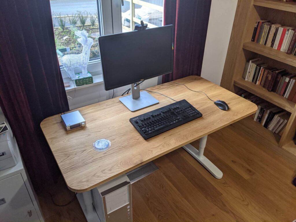 Modern home office with a wooden desk, monitor, keyboard, and bookshelf, with a view of a garden outside.