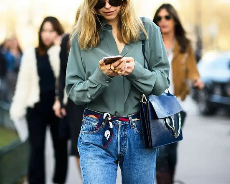 Stylish woman in sunglasses checks phone, wearing green blouse and jeans on a city street with a blue handbag.