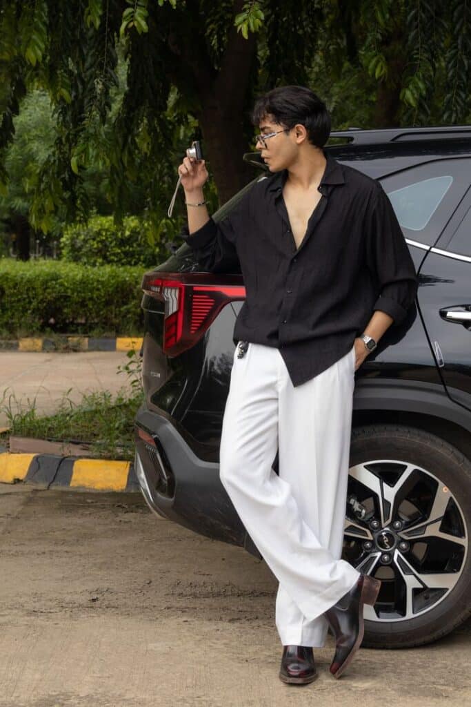Man in black shirt and white pants leaning on a car, taking a selfie in a scenic outdoor setting.
