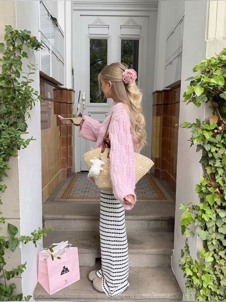 Woman in pink sweater with braided hair stands outside door, holding coffee and bag, surrounded by plants.