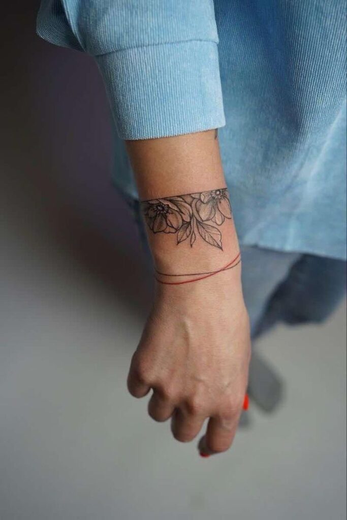 Floral wrist tattoo with red thread bracelet on forearm, person wearing a blue shirt.