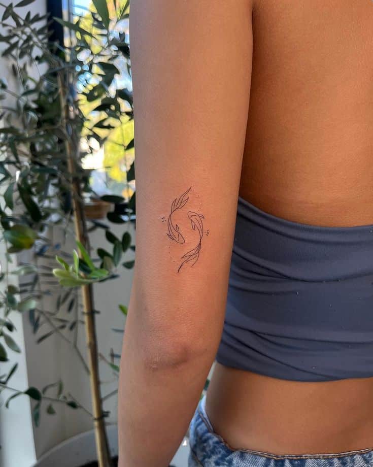 Arm tattoo with abstract fish design on a woman wearing a blue top, with plants in the background.