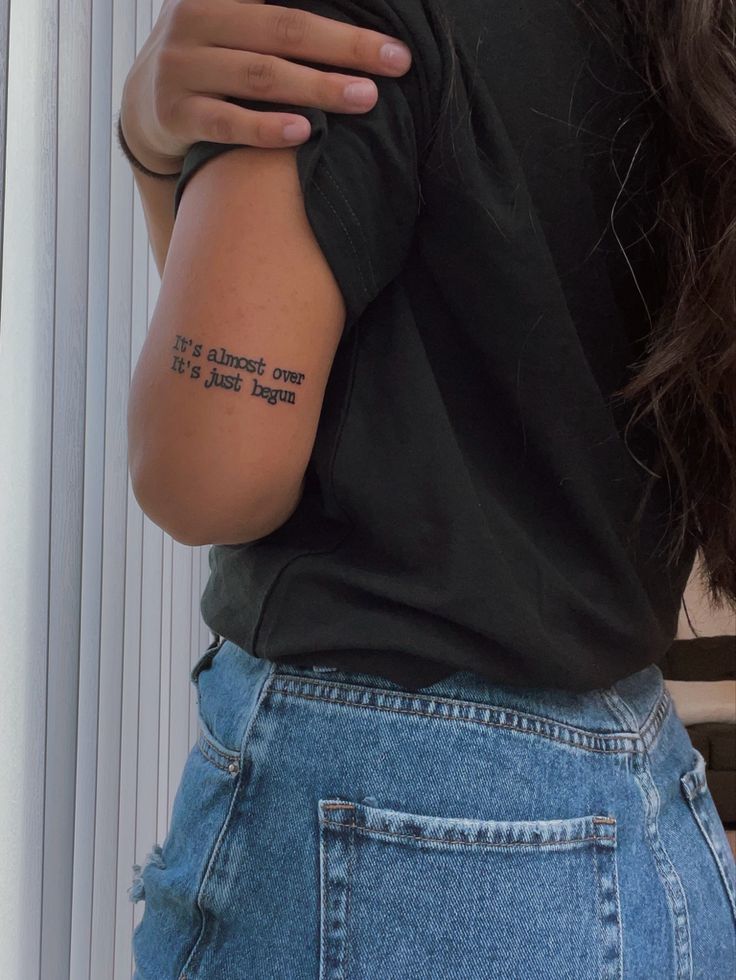 Person in black shirt with motivational tattoo text on arm, wearing blue jeans in natural light.