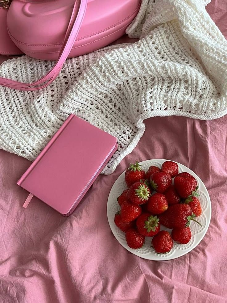 Pink purse, notebook, and strawberries on a white knit blanket and pink fabric. Cozy and stylish arrangement.