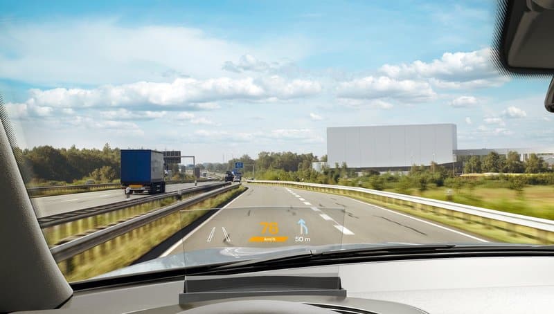 Car dashboard view with HUD speedometer on highway, showing 76 km/h and navigation directions under clear sky.