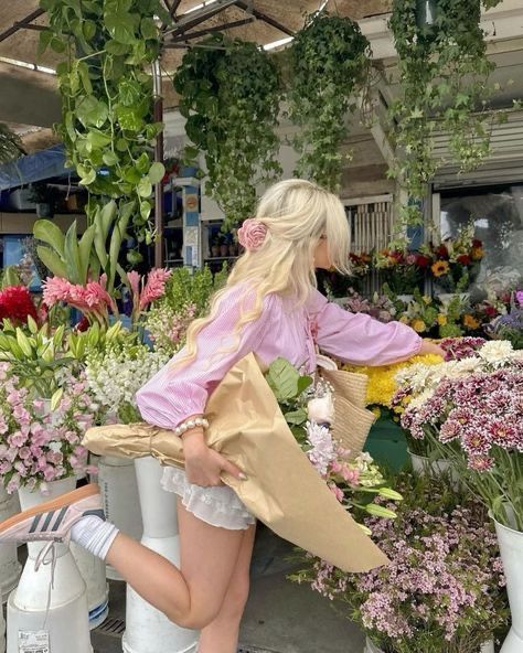Woman in pink blouse selecting flowers at a lush outdoor flower market, carrying a bouquet and basket.