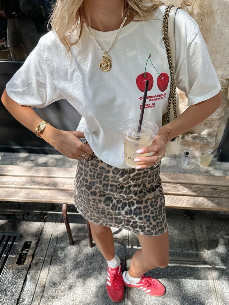 Woman wearing a cherry print t-shirt and leopard skirt, holding iced coffee. Fashionable street style with red sneakers.