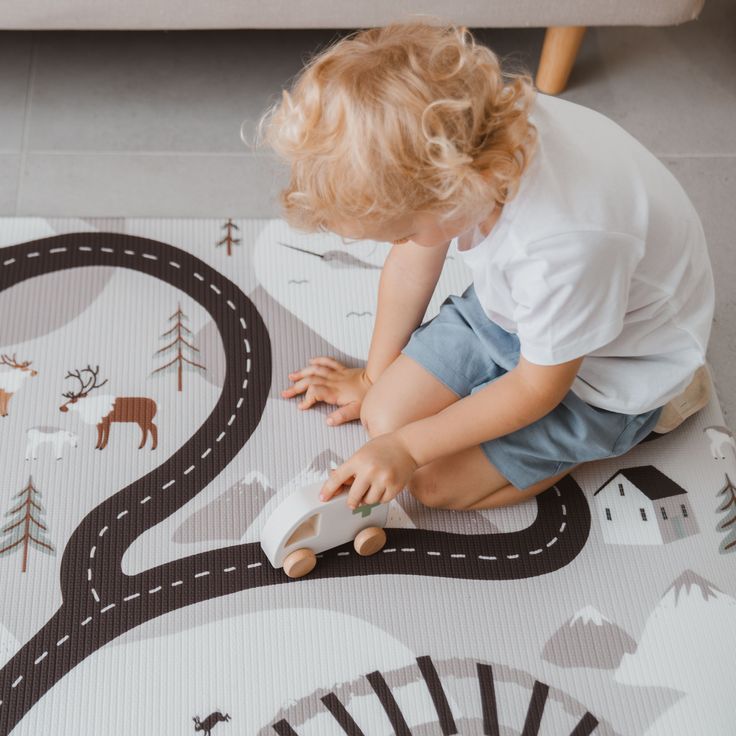 Child playing with toy car on a road-themed play mat, featuring trees and animals, in a cozy home setting.