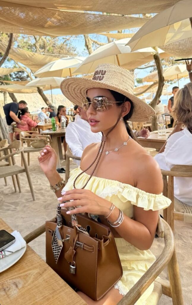 Woman in a sun hat and sunglasses dining outdoors, holding a stylish brown handbag, enjoying a sunny day.