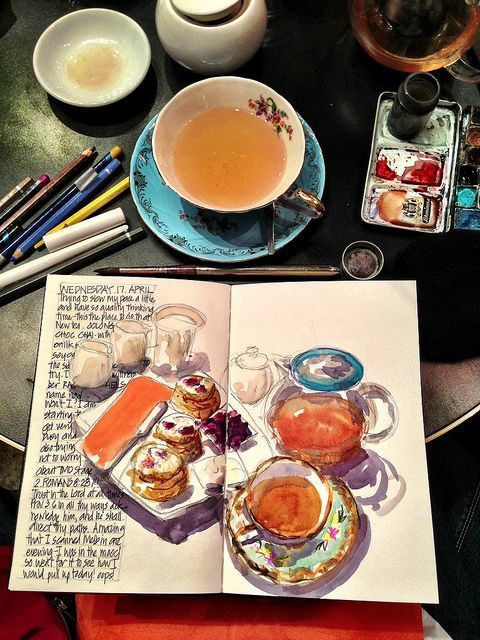 Sketchbook with tea and pastries illustration beside a cup of tea and art supplies on a table.