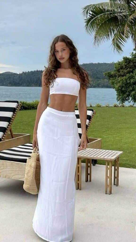 d45ef2f1-14e0-4604-b112-416209dd9e70 Woman in white summer outfit by the seaside, holding a straw bag, standing on a patio with palm trees in the background. | Sky Rye Design Woman in white summer outfit by the seaside, holding a straw bag, standing on a patio with palm trees in the background.
