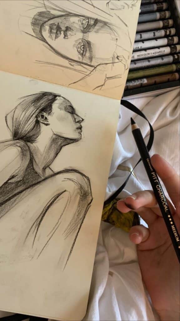 Charcoal sketches of a woman in a notebook with pencils nearby, artist holding a Pitt charcoal pencil.