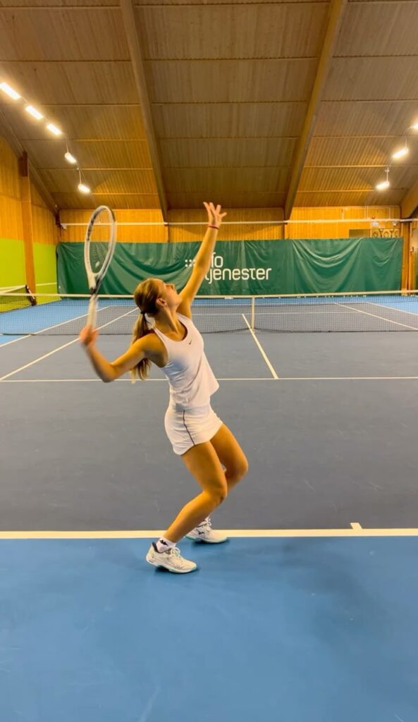 Tennis player in white outfit preparing to serve on an indoor court.