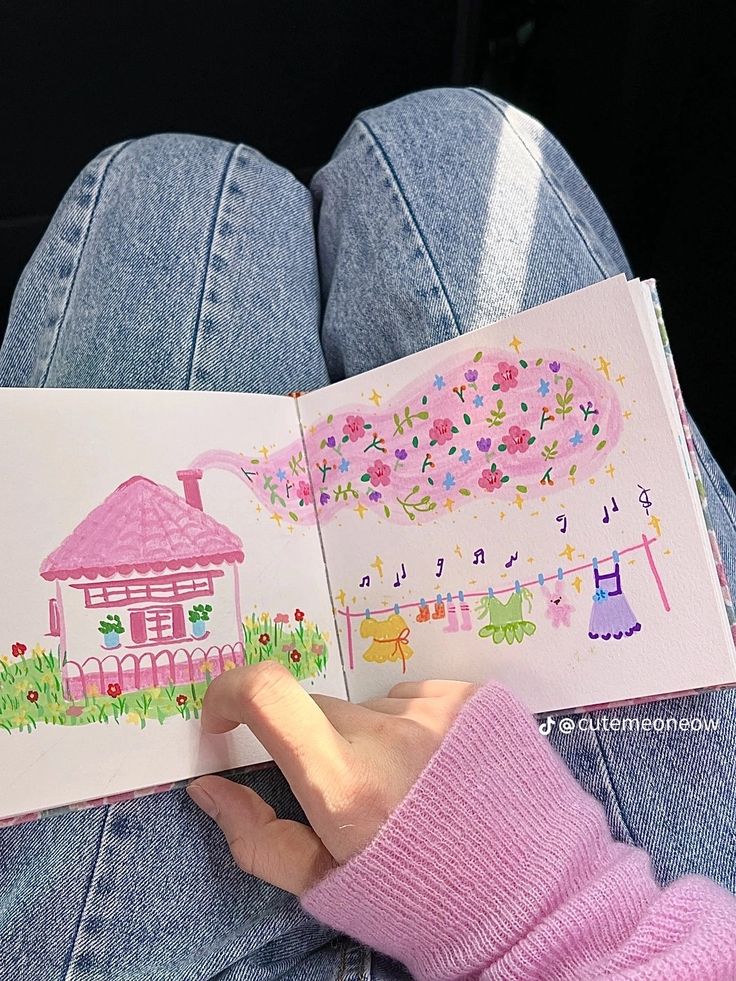 Hand holding an open sketchbook with colorful illustrations of a pink house and clothesline, on denim jeans background.