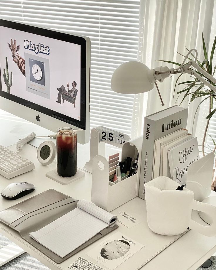 Modern workspace with desktop computer, office supplies, and a beverage on a white desk by a window with blinds.