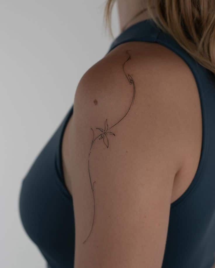 Minimalist floral tattoo on woman's shoulder, wearing a dark tank top, showcasing elegant and delicate line art design.