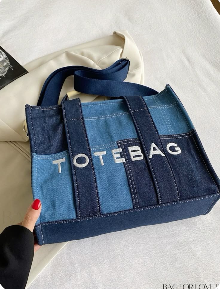 Denim tote bag with blue straps and TOTE BAG text, on a cream background, held by a person with red nails.