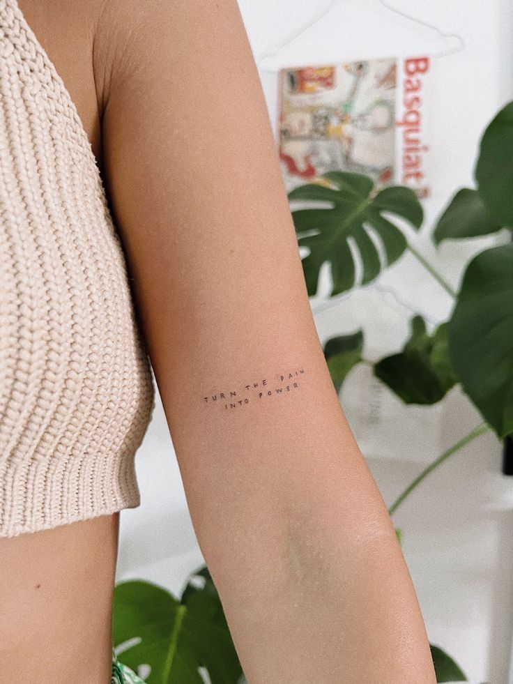 Minimalist arm tattoo reading Turn the pain into power on person in knitted top with green plant background.