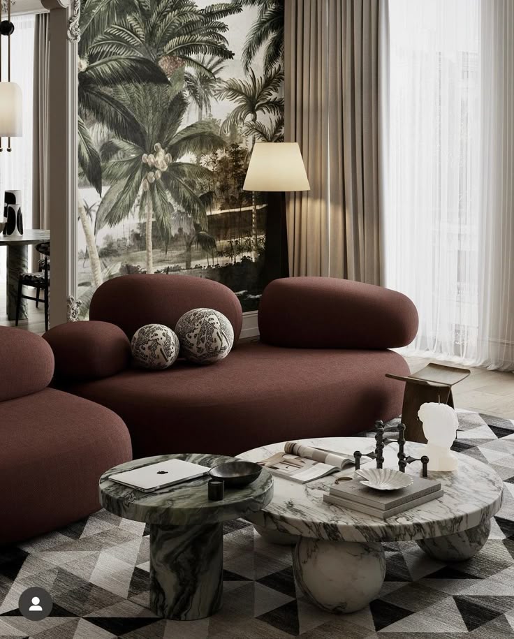 Modern living room with maroon sofa, tropical wallpaper, and marble coffee tables in natural light.