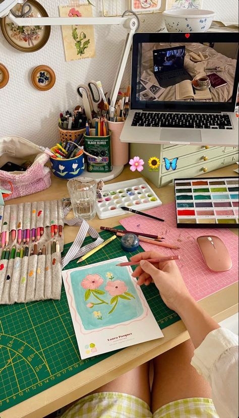 Cozy art workspace with paints, brushes, and laptop, featuring a floral watercolor painting in progress.