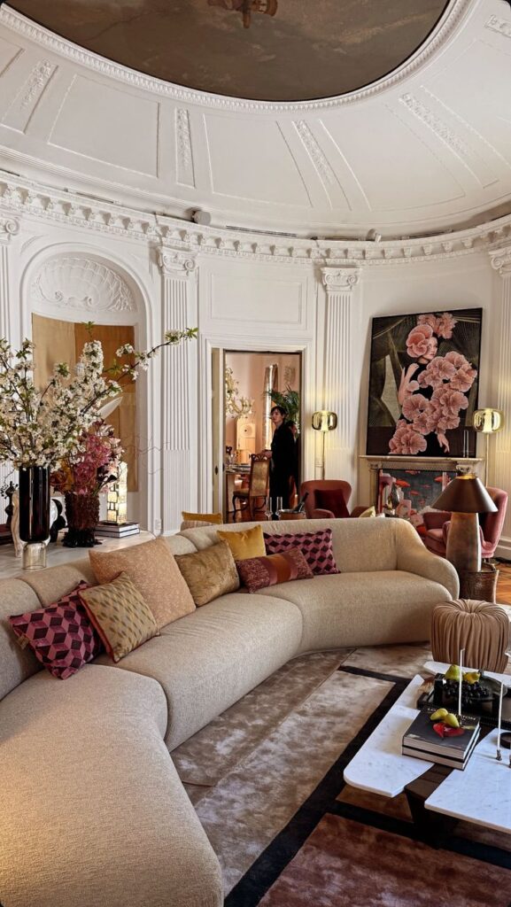 Elegant living room with curved beige sofa, colorful pillows, floral decor, and ornate ceiling details.
