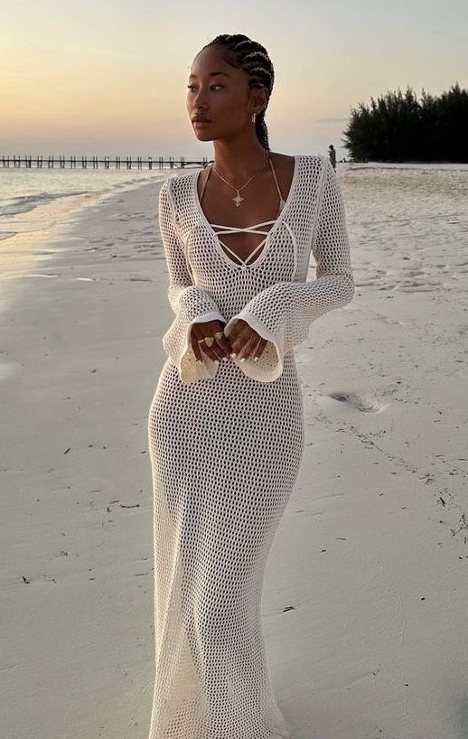Woman in a white crochet dress on a beach at sunset, showcasing elegant fashion and serene coastal vibes.