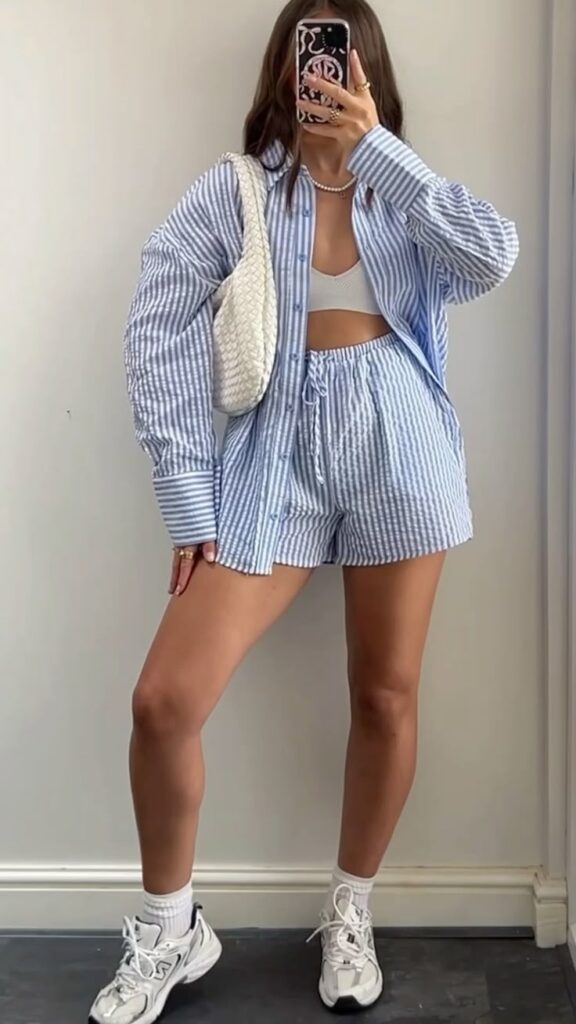 Woman in blue striped outfit with white sneakers and bag, taking a mirror selfie indoors. Casual summer style.