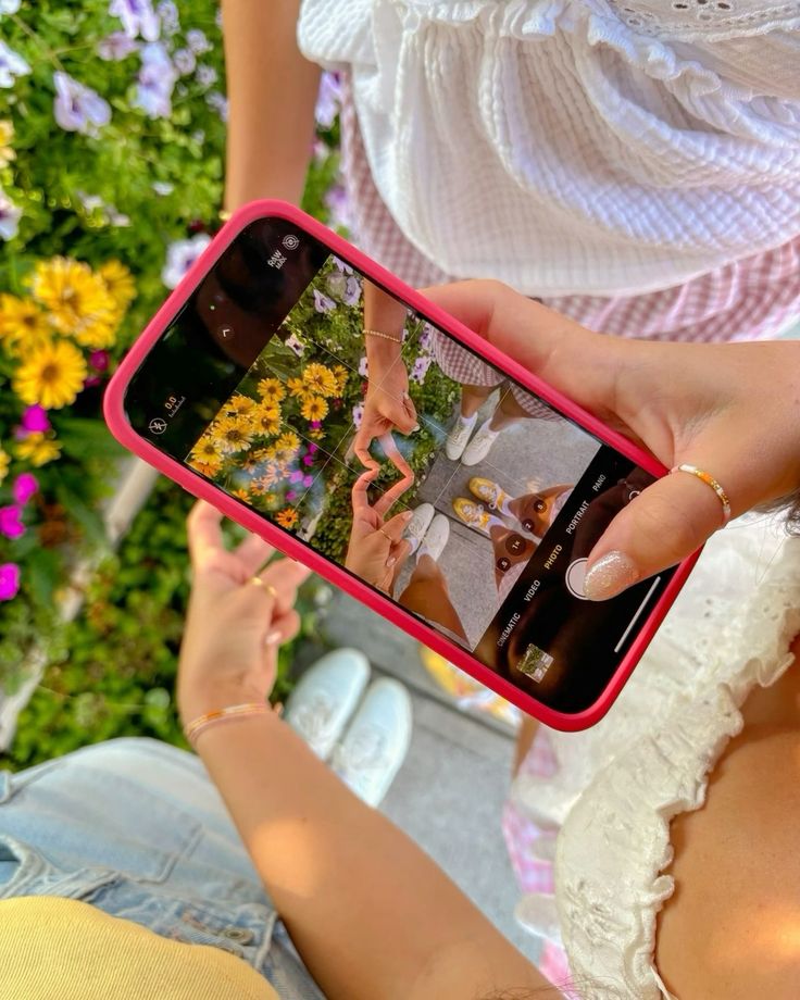 Capturing a heart shape on smartphone near vibrant flowers, with friends enjoying a sunny day.