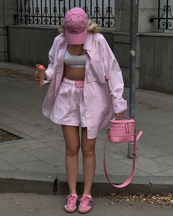 Woman in pink striped outfit with matching cap and shoes, holding ice cream and pink bag on city sidewalk.