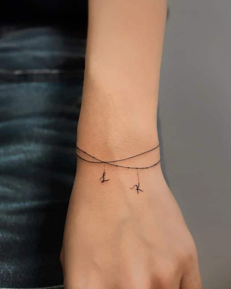 Minimalistic wrist tattoo featuring delicate strings with small cursive letters, resembling jewelry.