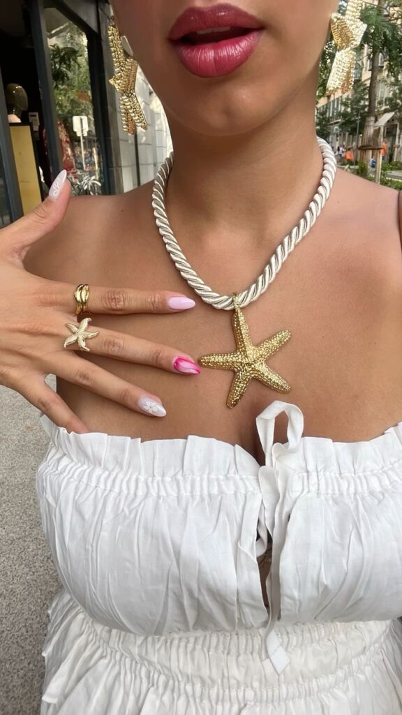 Woman in white dress with gold starfish jewelry, pink lipstick, and pastel nail art outdoors.