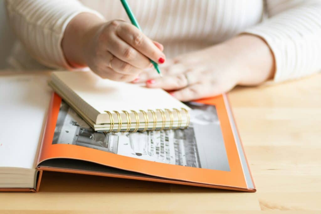 Person writing in notebook on wooden desk, next to an open photo album.