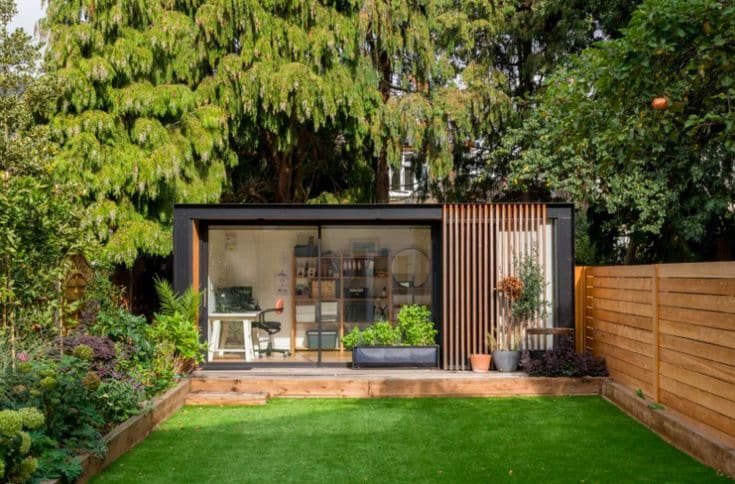 Modern garden office with glass front, surrounded by lush greenery and wooden fencing.