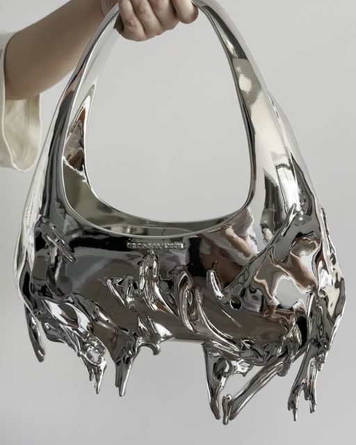 Futuristic chrome handbag with abstract design held in woman's hand against a plain background.