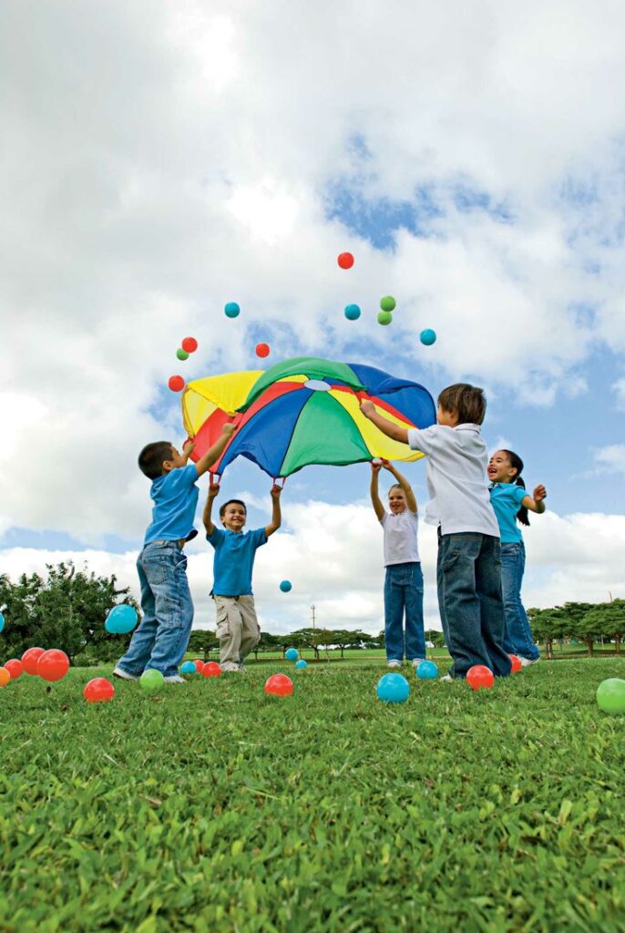 Children playing with a colorful parachute and balls on a grassy field under a clear blue sky.