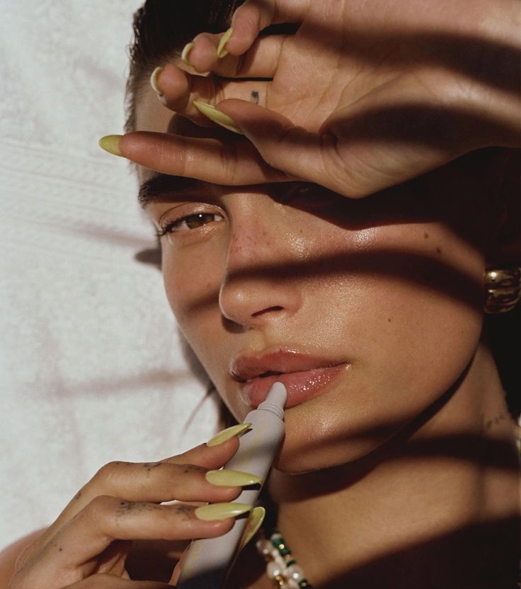 Woman applying lip balm in sunlight, with hand shading face. Natural makeup, long nails, and sunlit beauty.