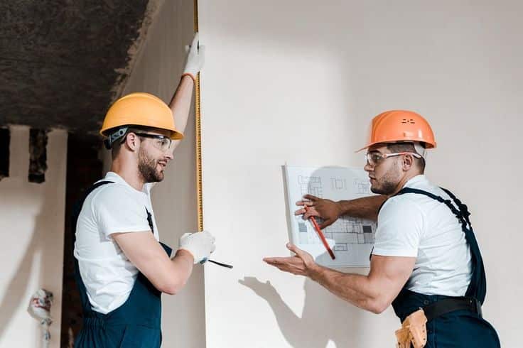 Two construction workers measuring and discussing building plans on a site, wearing helmets and safety gear.