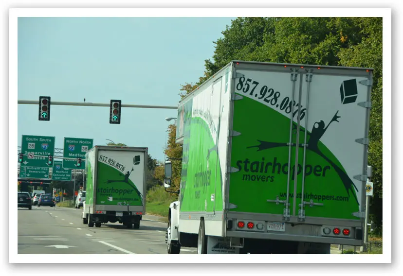 Two green Stairhopper Movers trucks driving on a highway under a green traffic light near directional signs.